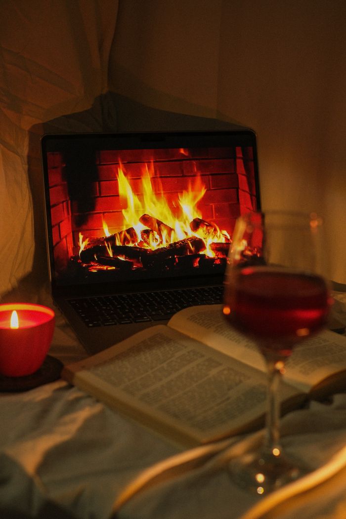 Home A serene setup with a laptop fireplace, open book, red wine, and a candle, epitomizing cozy indoor relaxation.